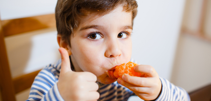 Young boy eating an orange and doing a thumbs up to the camera