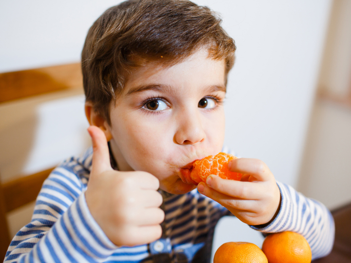 Young boy eating an orange and doing a thumbs up to the camera