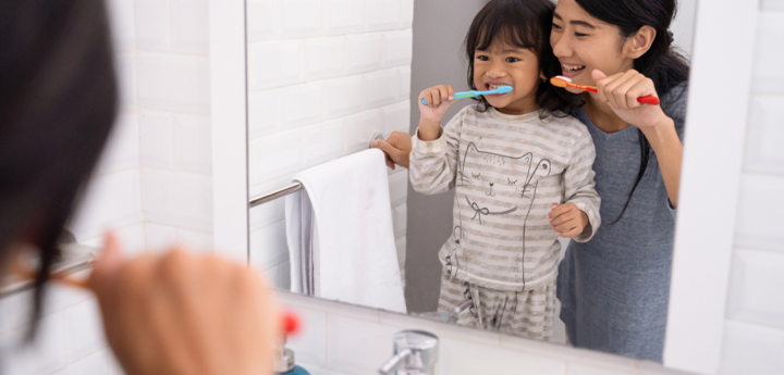 Young girl brushing their teeth with their mum looking into the bathroom mirror